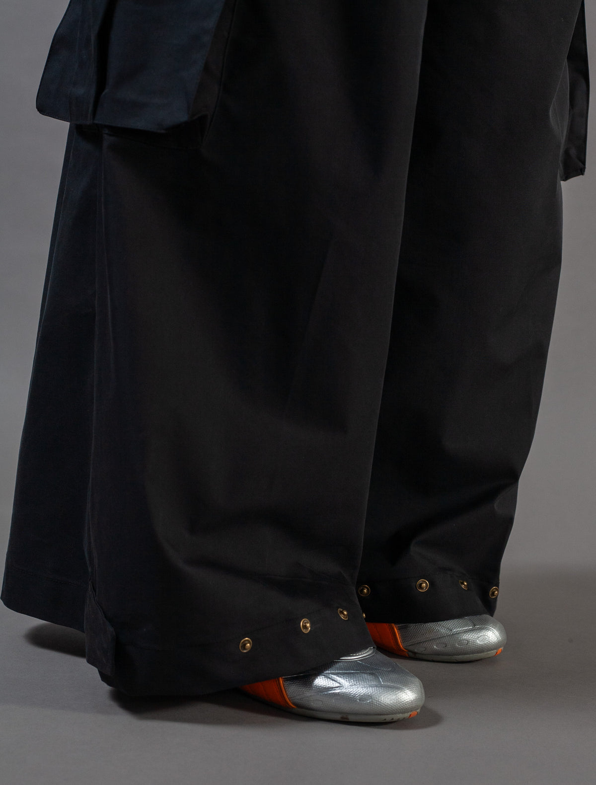 Person wearing a long black pants with metallic shoes on a gray background