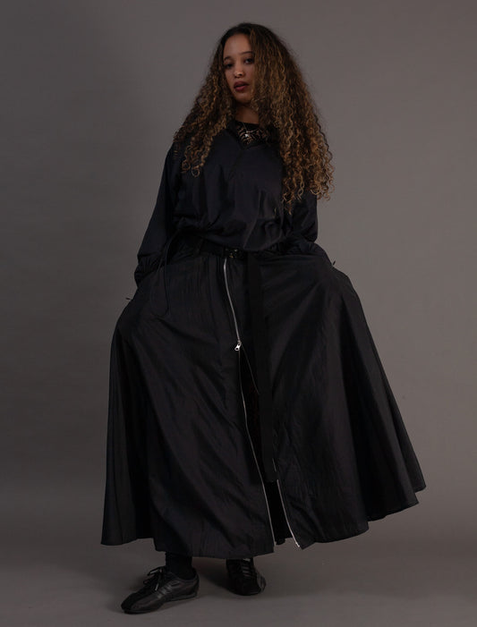 Person wearing a black outfit with a long skirt and jacket on a gray background