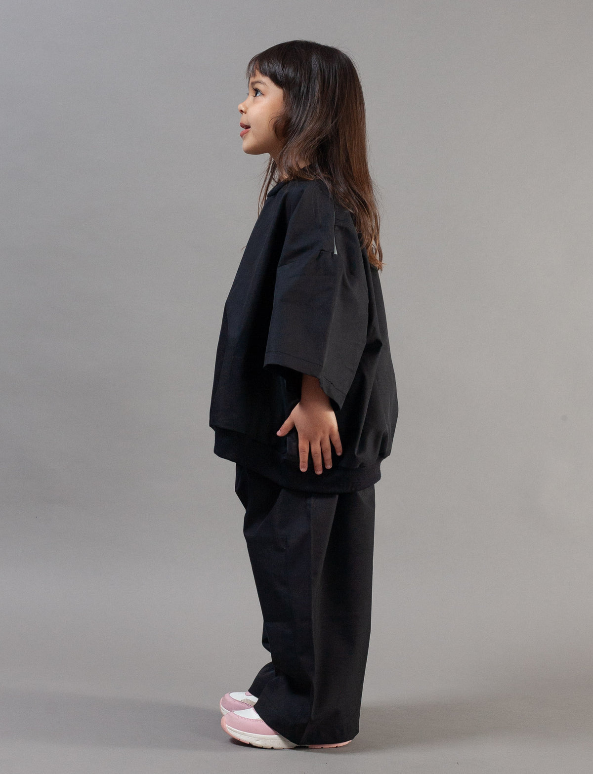 A young girl with long dark hair is shown in profile wearing a black wide-sleeved top and matching black pants, standing against a solid gray studio background.