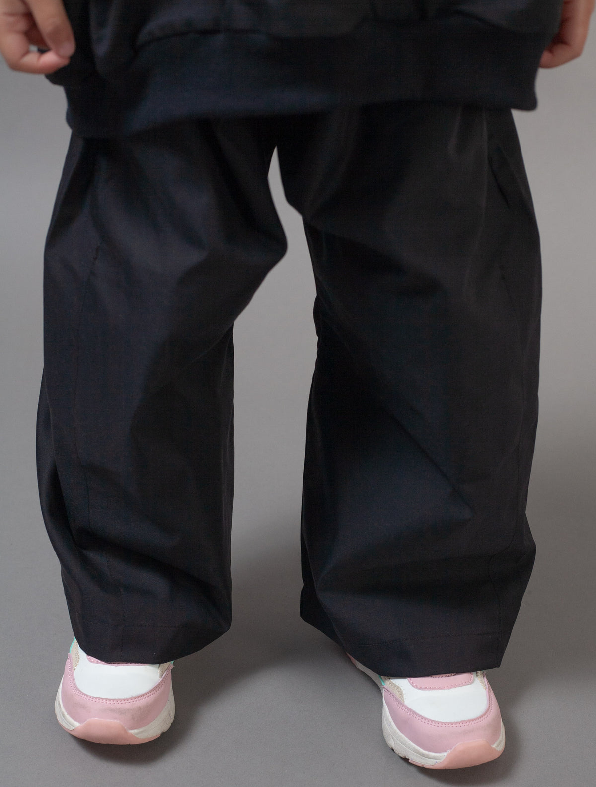 A close-up of a person's lower body wearing loose, oversized black pants and light pink and white sneakers against a solid gray studio background.
