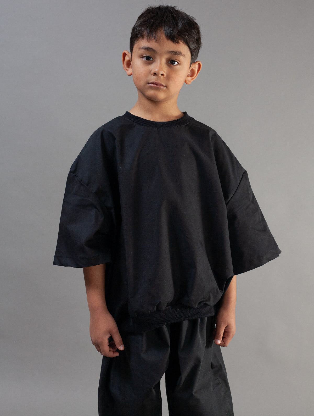A young boy wearing an oversized black short-sleeve top and matching black pants, standing against a plain gray studio background.