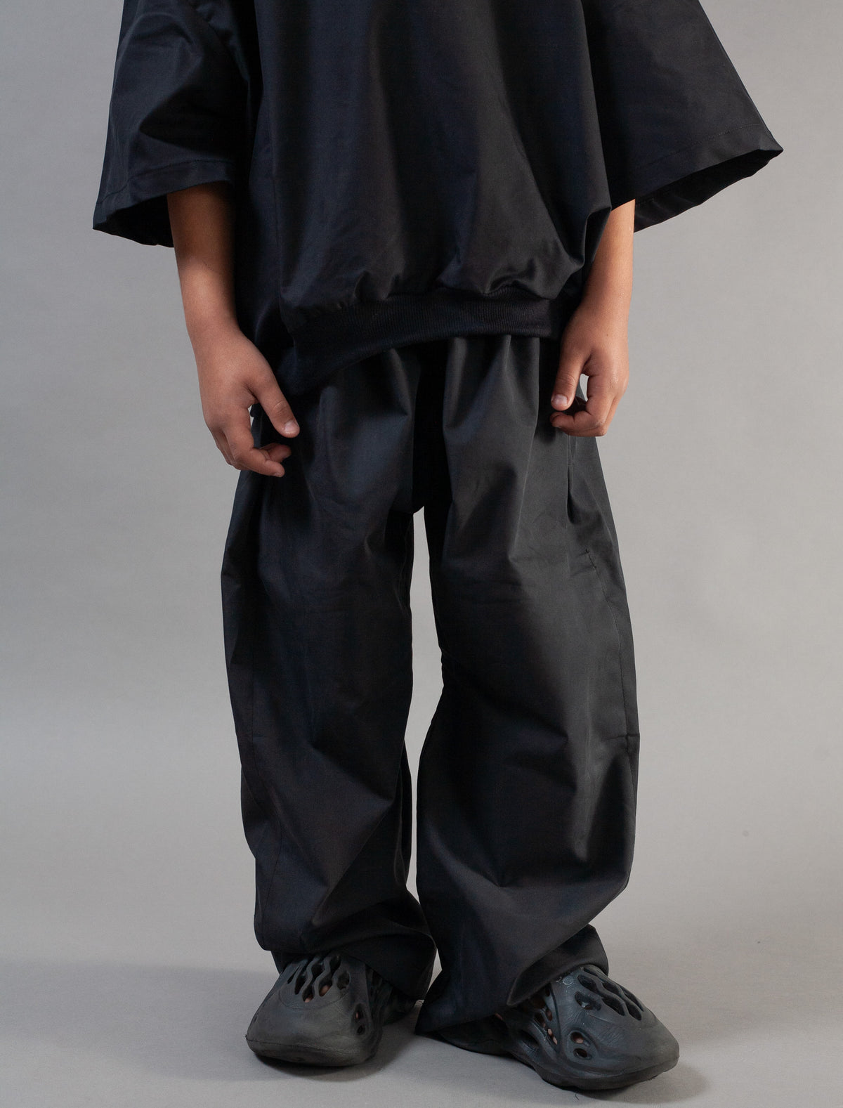 A person wearing loose black wide-leg pants and a matching oversized black top, standing against a plain light gray studio background.