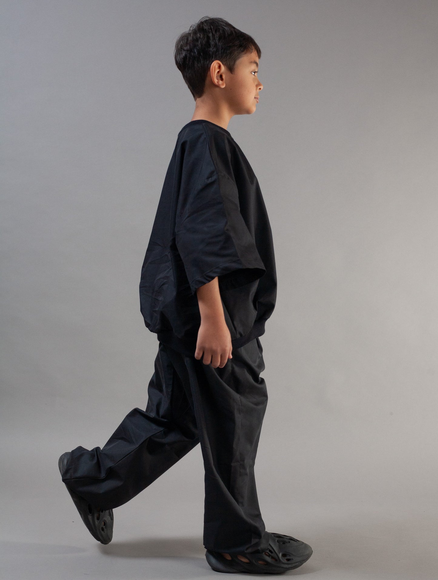 A child model in profile wearing a loose black oversized top and matching black wide-leg pants, photographed against a plain light gray studio background.