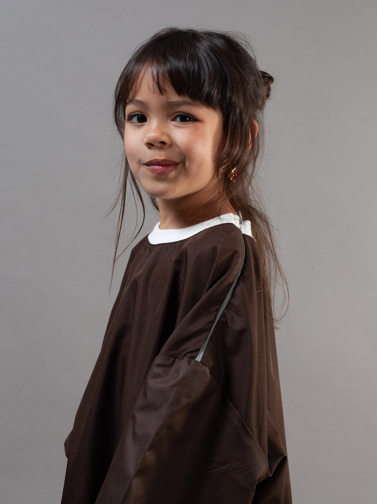 A young girl with dark hair wearing a brown salon cape over a white t-shirt, photographed against a solid gray studio background.