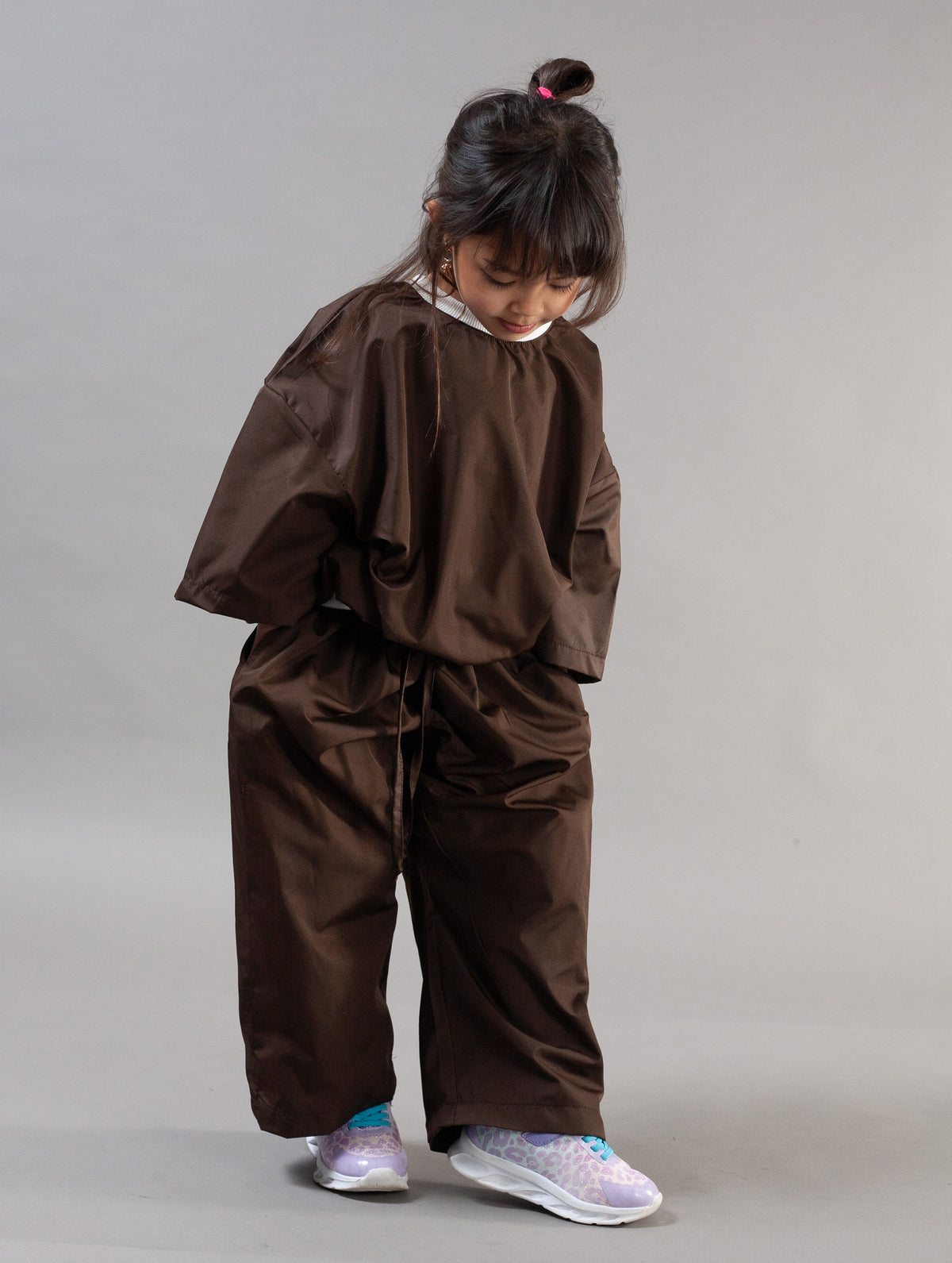 A child wearing an oversized brown hooded sweatshirt and matching loose brown pants, standing against a plain light gray studio background.