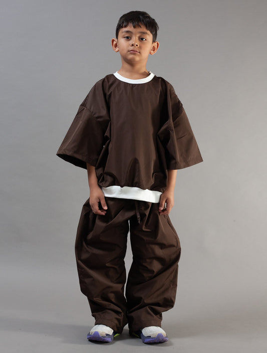 A young boy modeling a dark brown traditional Japanese-style outfit with wide sleeves and loose pants, featuring white contrast trim at the collar and hem, against a solid gray studio background.