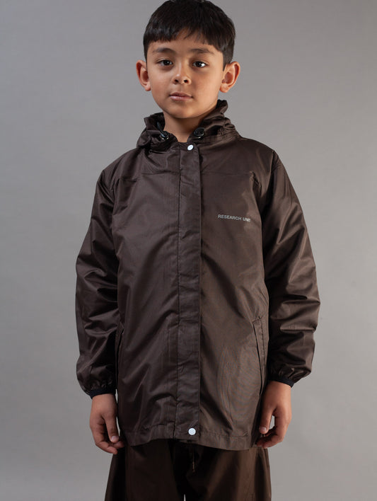 A young boy wearing a dark brown waterproof hooded jacket with a small white brand logo on the left chest, photographed against a neutral gray studio background.