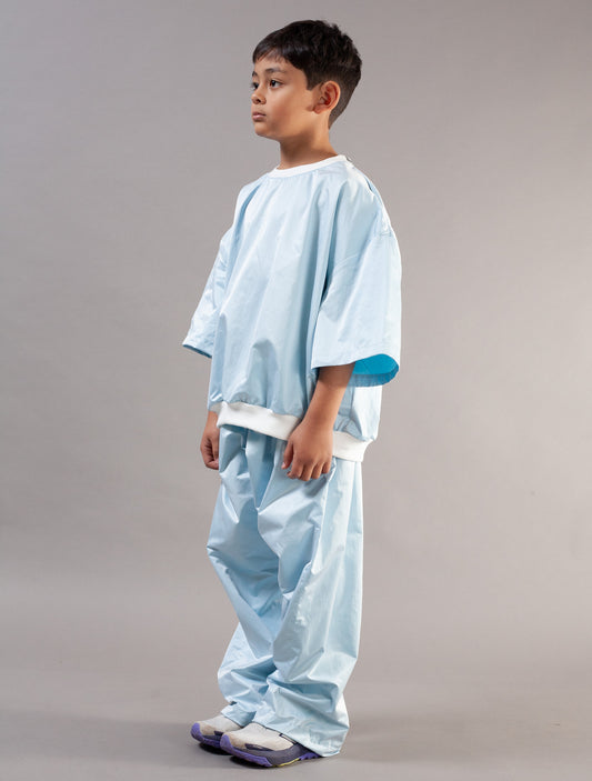 A young boy modeling a light blue kids set, consisting of a short-sleeve top and matching pants, standing against a neutral gray studio background.