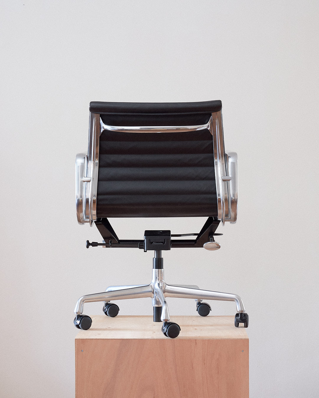 A black mesh office chair with a chrome five-star base and casters, photographed from the back and resting on a light wood block against a plain off-white wall.