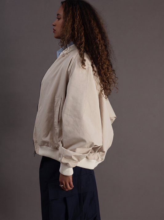 A woman shown in side profile wearing a cream-colored oversized bomber jacket with voluminous sleeves and dark navy pants, set against a solid dark gray studio background.