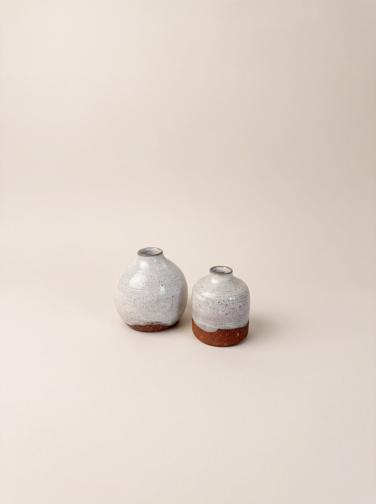 Two small ceramic vases with rounded bulbous bodies, speckled light gray glazes, and exposed terracotta bases, photographed on a plain warm beige studio background.
