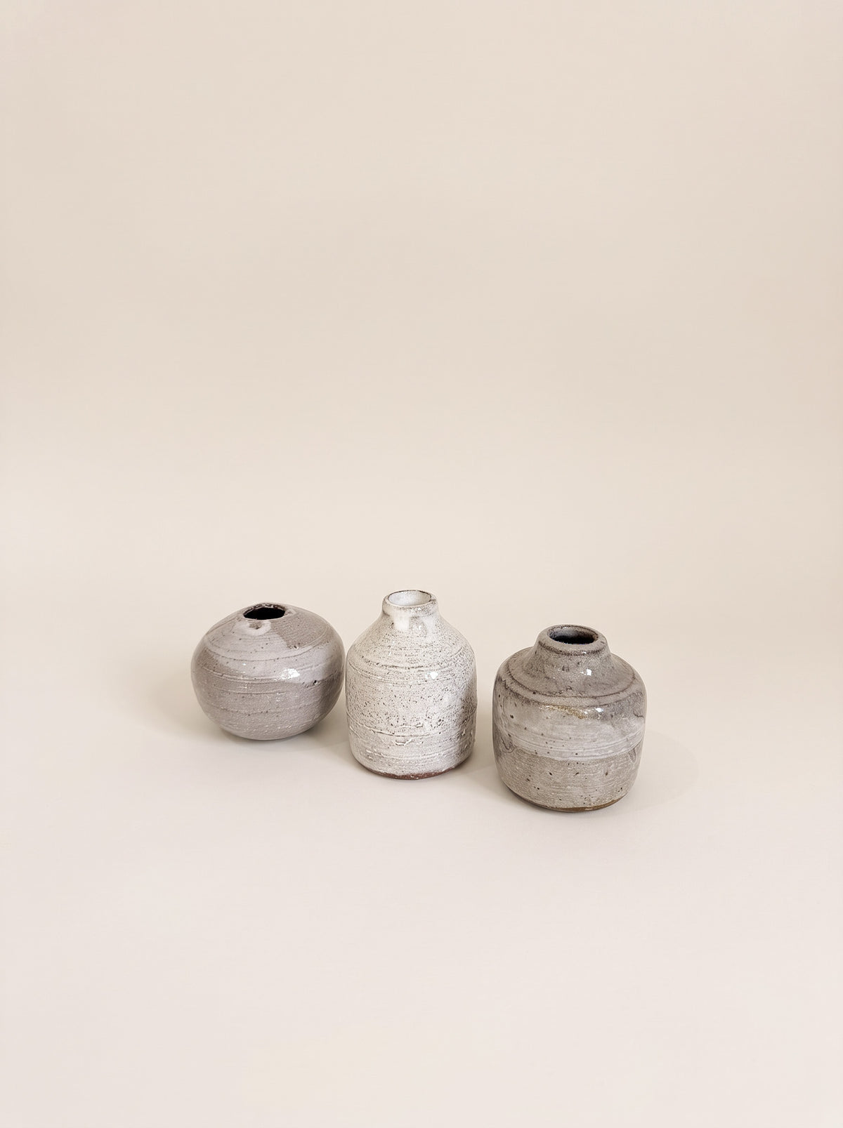 Three small handmade ceramic vases in textured gray and white glazes, arranged in a row against a solid warm beige background.