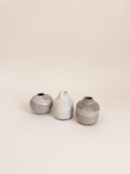 Three small handmade ceramic vases in textured gray and white glazes, arranged in a row against a solid warm beige background.