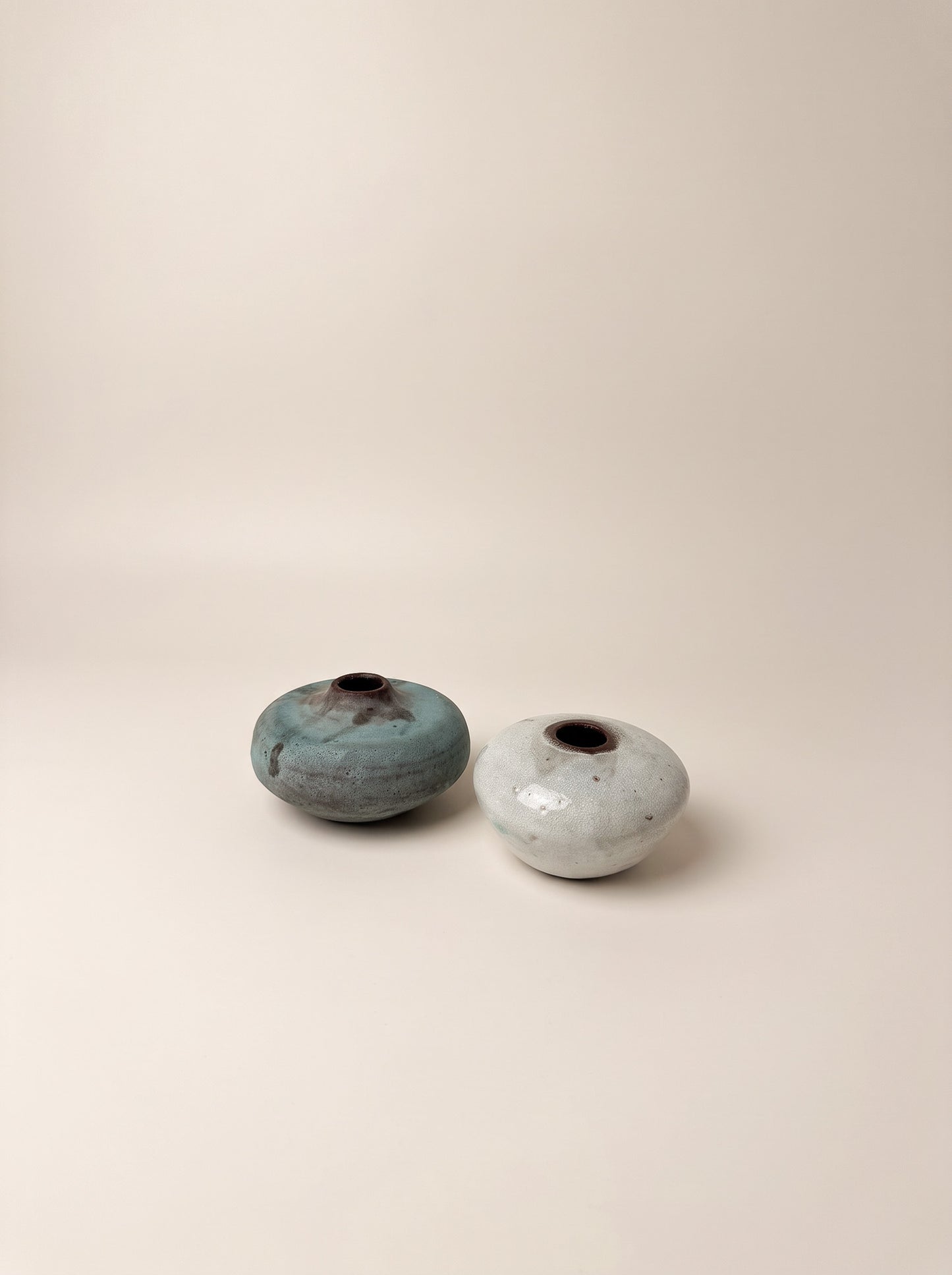 Two small handmade ceramic vases with rounded, egg-shaped bodies and small dark openings, displayed on a warm beige studio background. One vase has a textured teal-blue glaze with brown speckling, and the other has a speckled off-white glaze.
