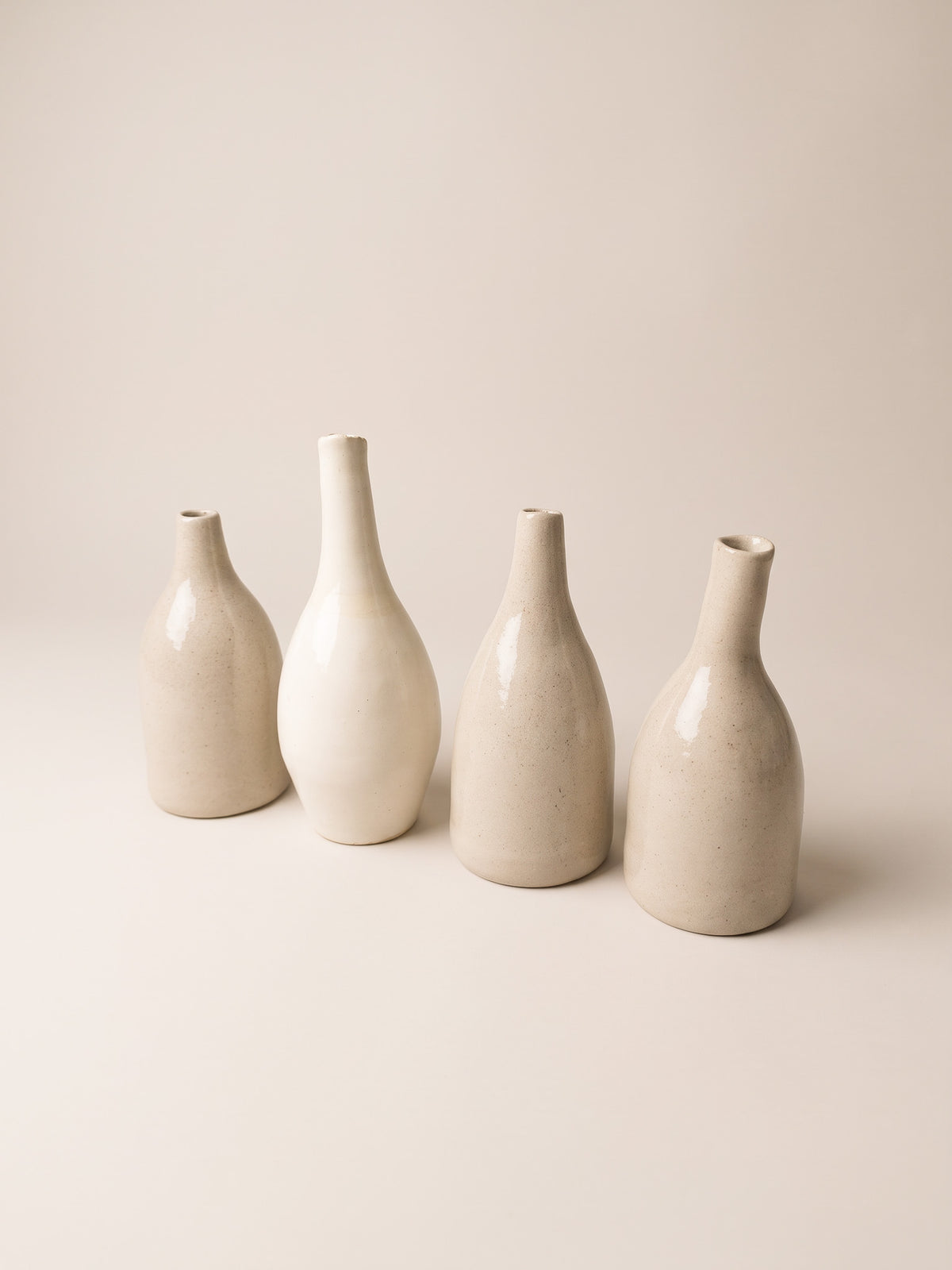 Four minimalist ceramic vases in warm off-white and cream tones, arranged in a row against a solid, light beige background.