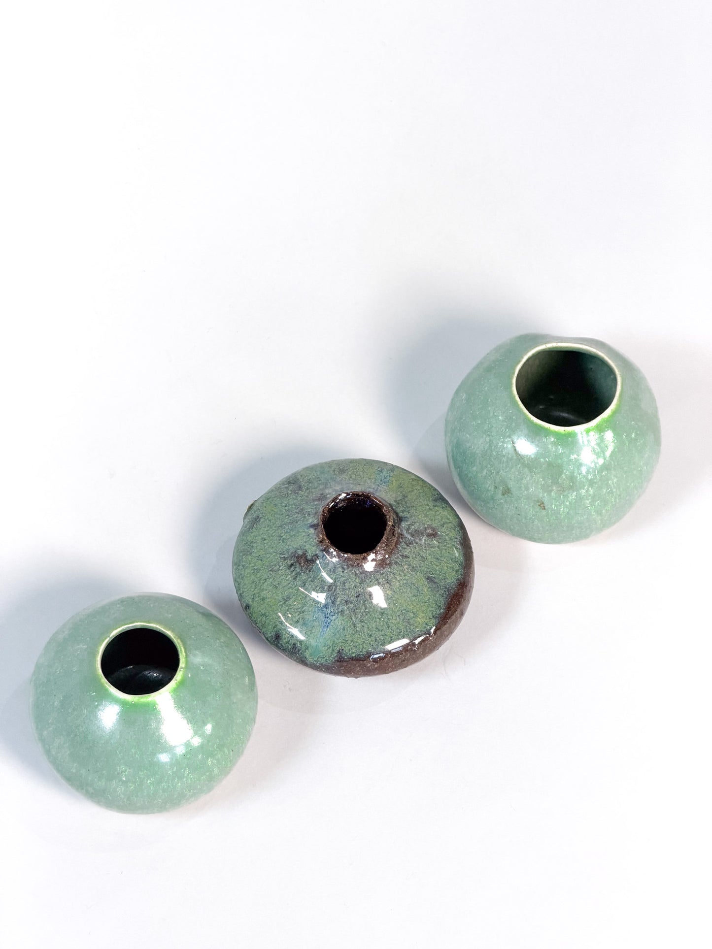 Three small round ceramic vases with a glossy light green glaze and dark centers, arranged diagonally on a plain white background.