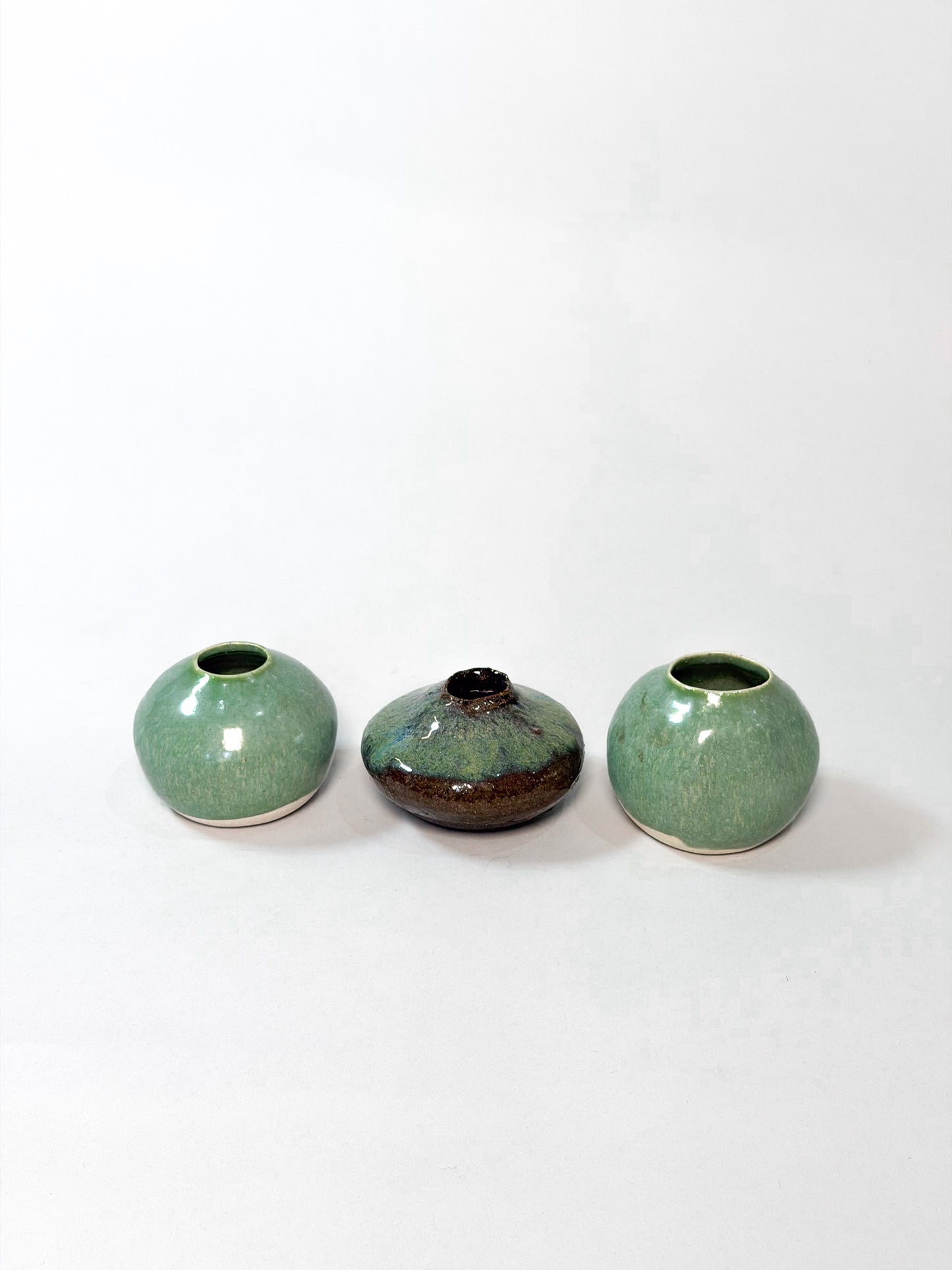 Three small ceramic vases arranged in a row on a plain white background. The two outer vases are rounded and glazed in a light green glaze, while the center vase is darker with a brown and black mottled finish.