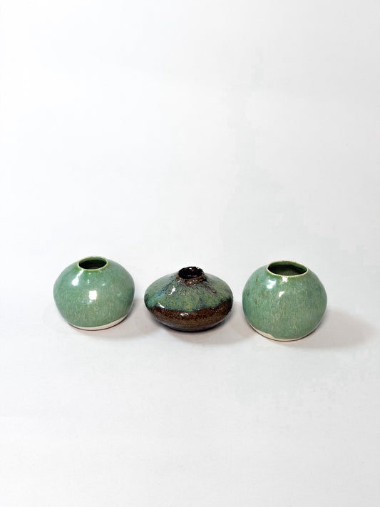 Three small ceramic vases arranged in a row on a plain white background. The two outer vases are rounded and glazed in a light green glaze, while the center vase is darker with a brown and black mottled finish.