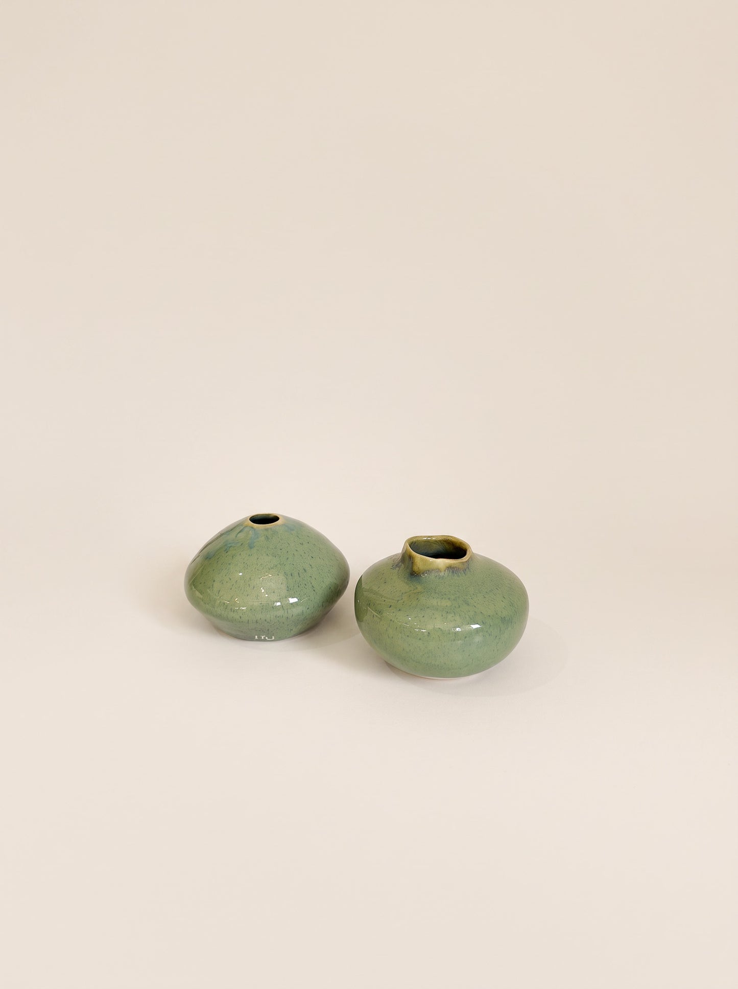 Two small rounded ceramic vases with a glossy green glaze and subtle gold-toned highlights, shown on a plain warm beige background.