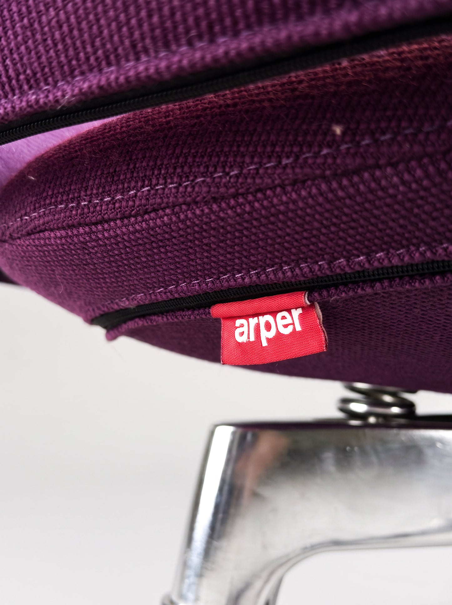 Close-up of a purple woven fabric product with a black zipper and a red Arper brand tag, shown against a white background with part of a metallic silver object visible below.
