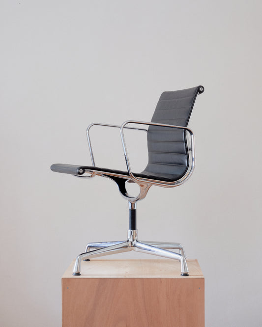 A mid-century modern armchair with a black ribbed vinyl seat and backrest, supported by a polished chrome five-star base and tubular frame, shown sitting on a light wood pedestal against a neutral off-white background.