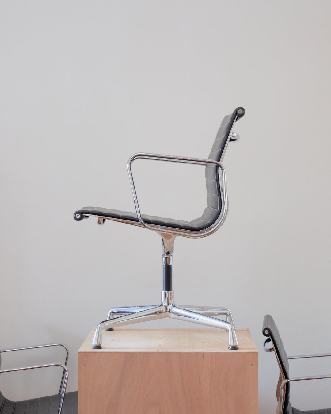 A mid-century modern armchair with a chrome metal frame and gray upholstered seat and backrest, displayed on a light wood pedestal against a plain off-white wall.