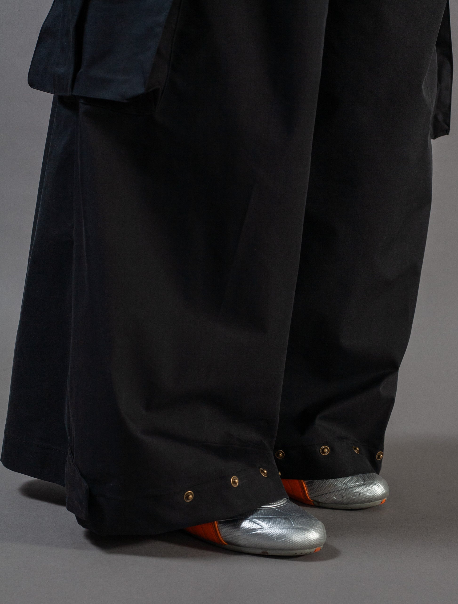 Person wearing a long black pants with metallic shoes on a gray background