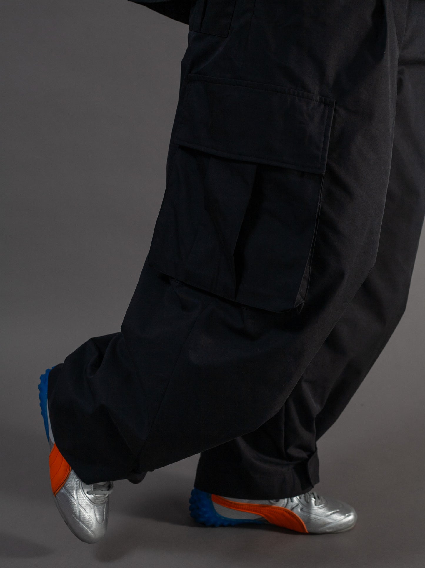 Person wearing black pants and silver shoes with orange soles on a gray background