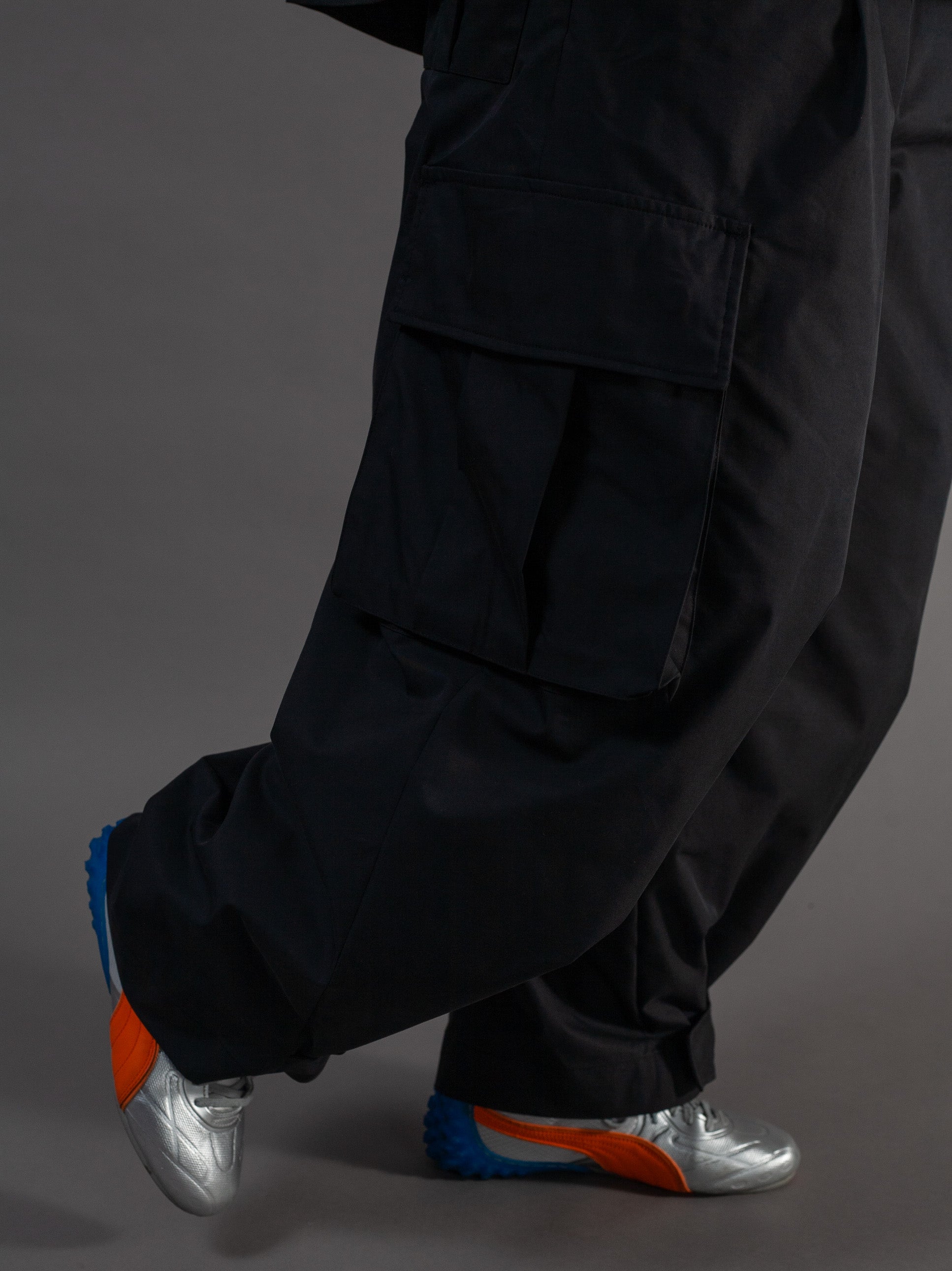 Person wearing black pants and silver shoes with orange soles on a gray background
