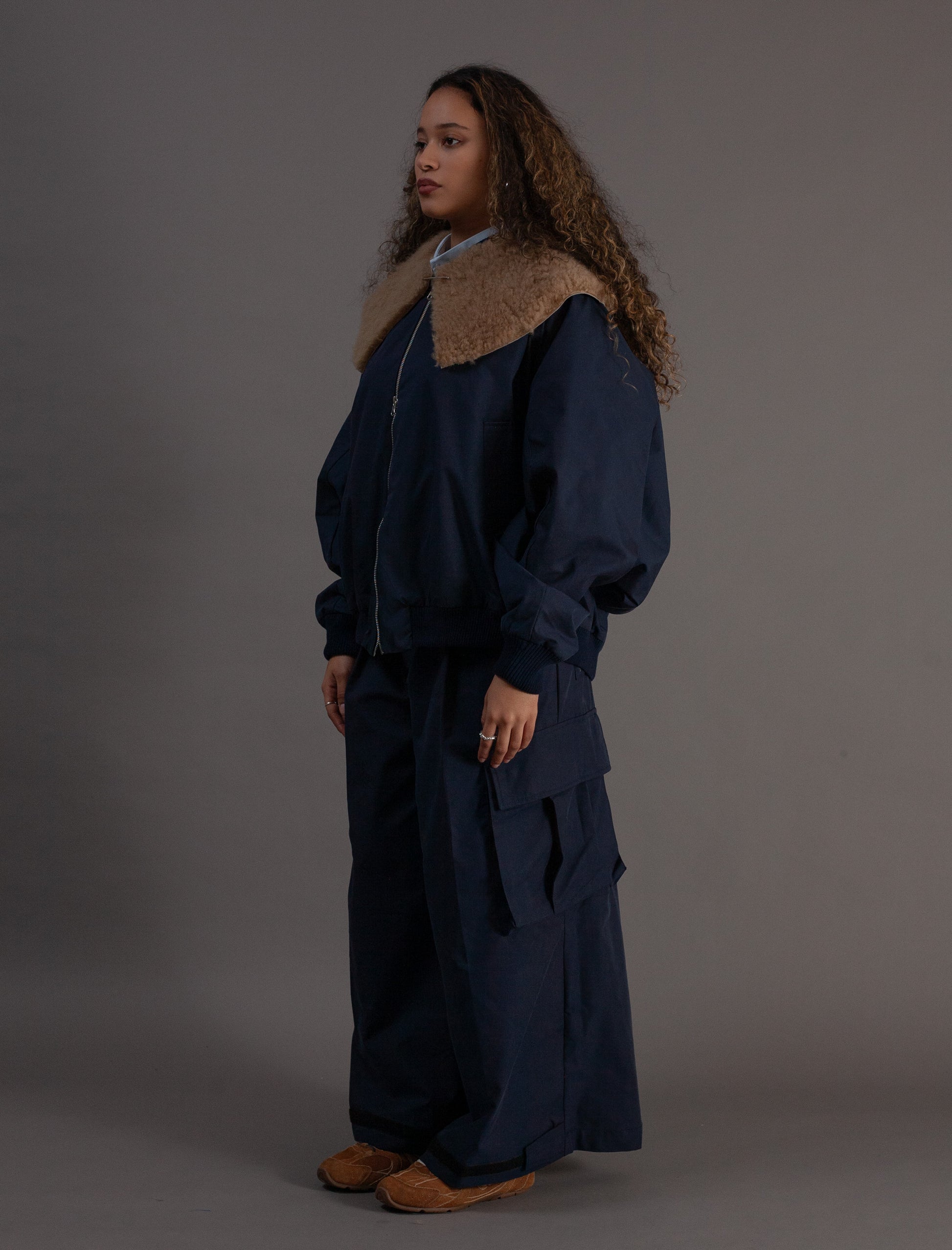 Person wearing a dark blue outfit with a fur-lined hood against a gray background