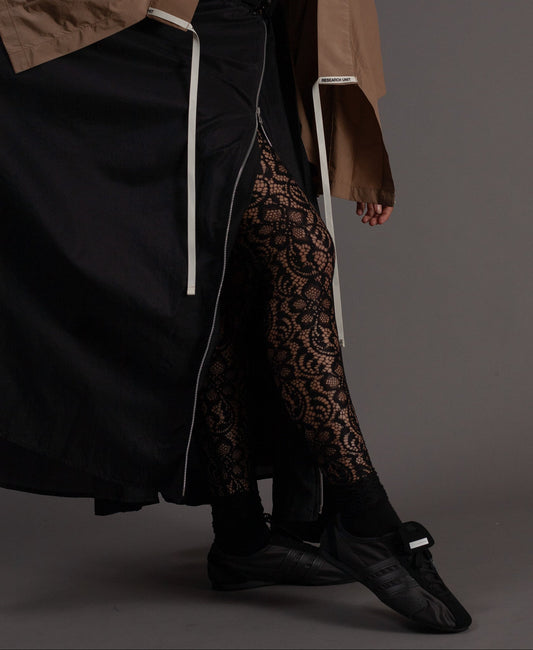 Person wearing a brown coat over a black dress with lace details on a gray background