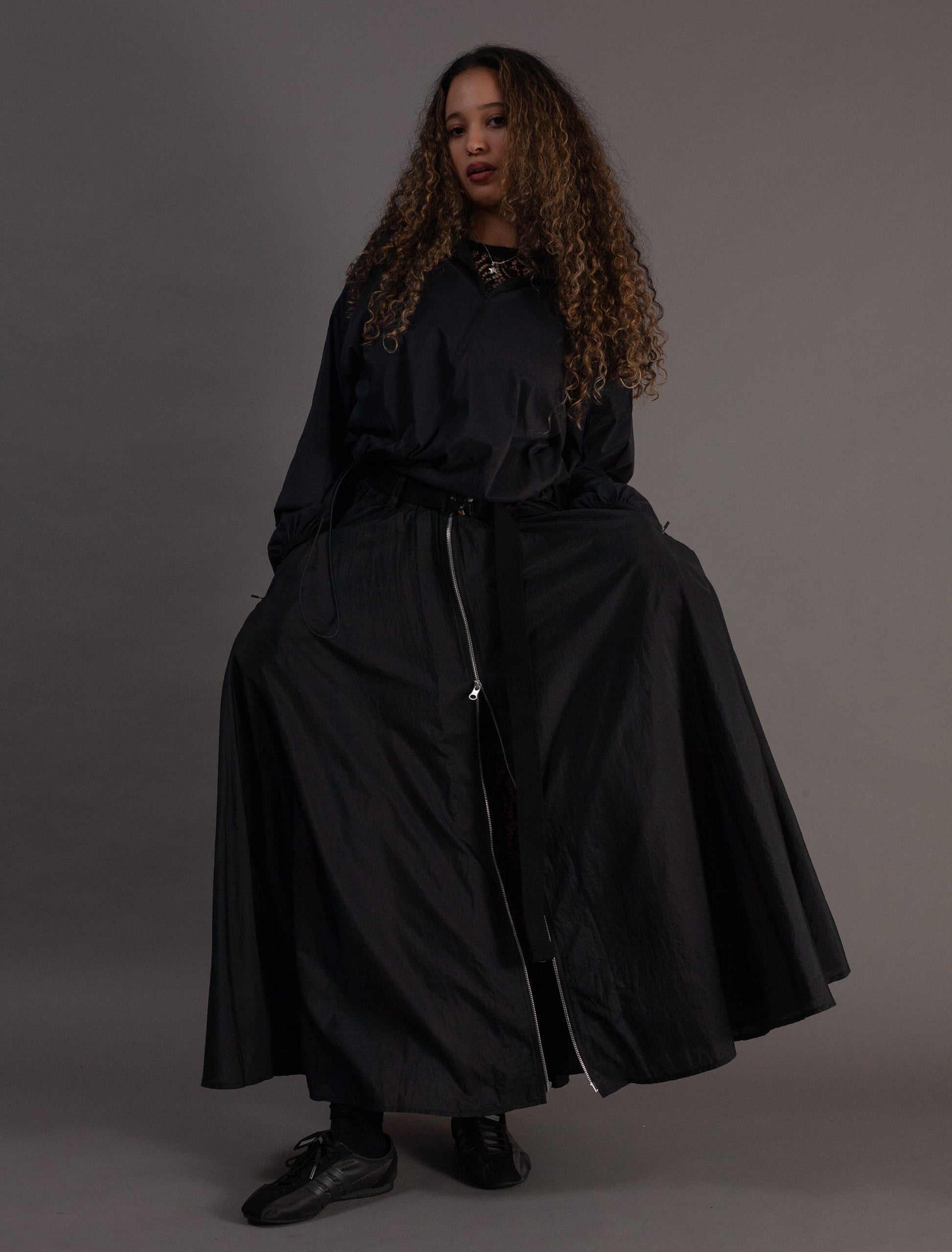 Person wearing a black outfit with a long skirt and jacket on a gray background
