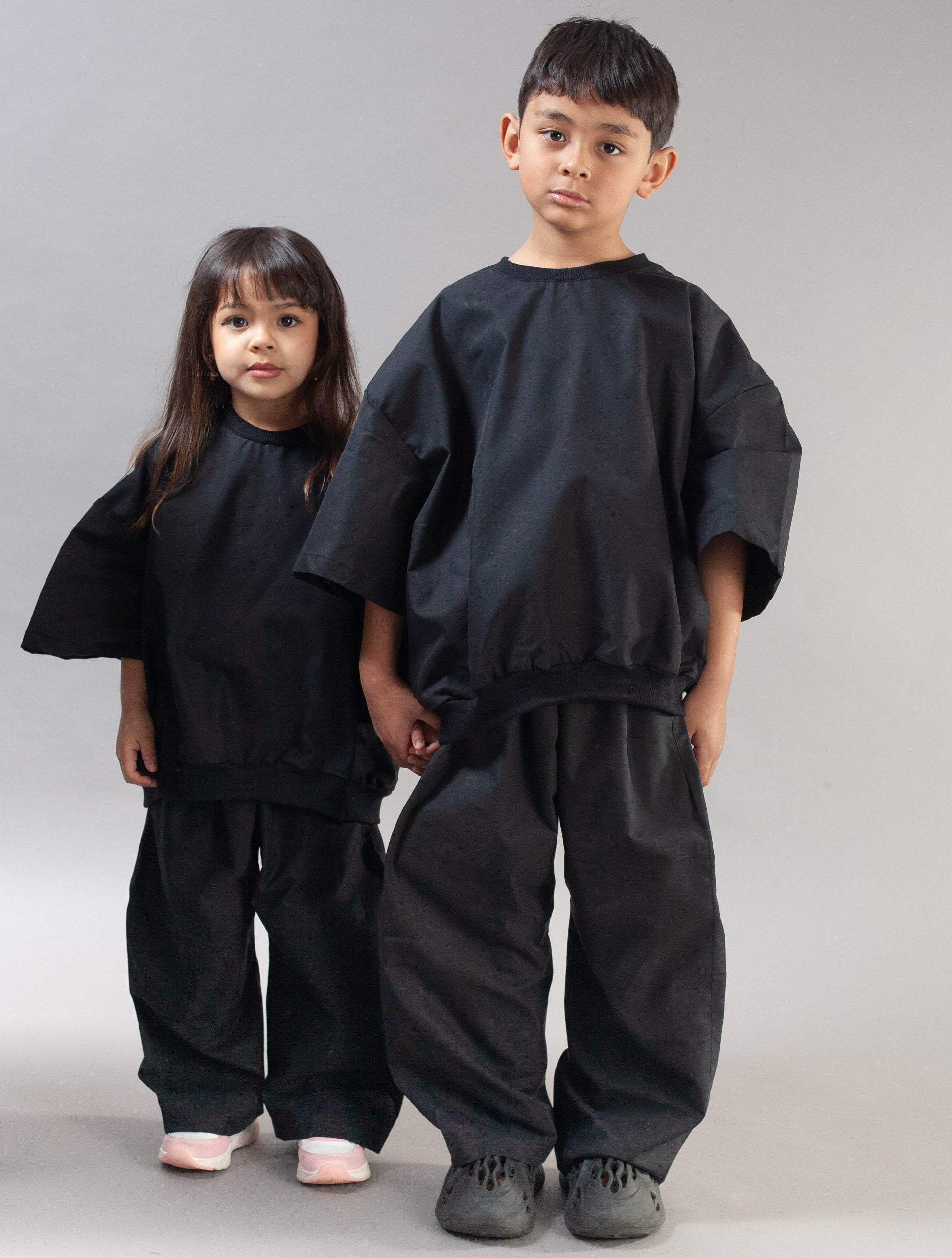 A young boy and a girl modeling matching black wide-sleeve, loose-fitting outfits against a plain light gray studio background.