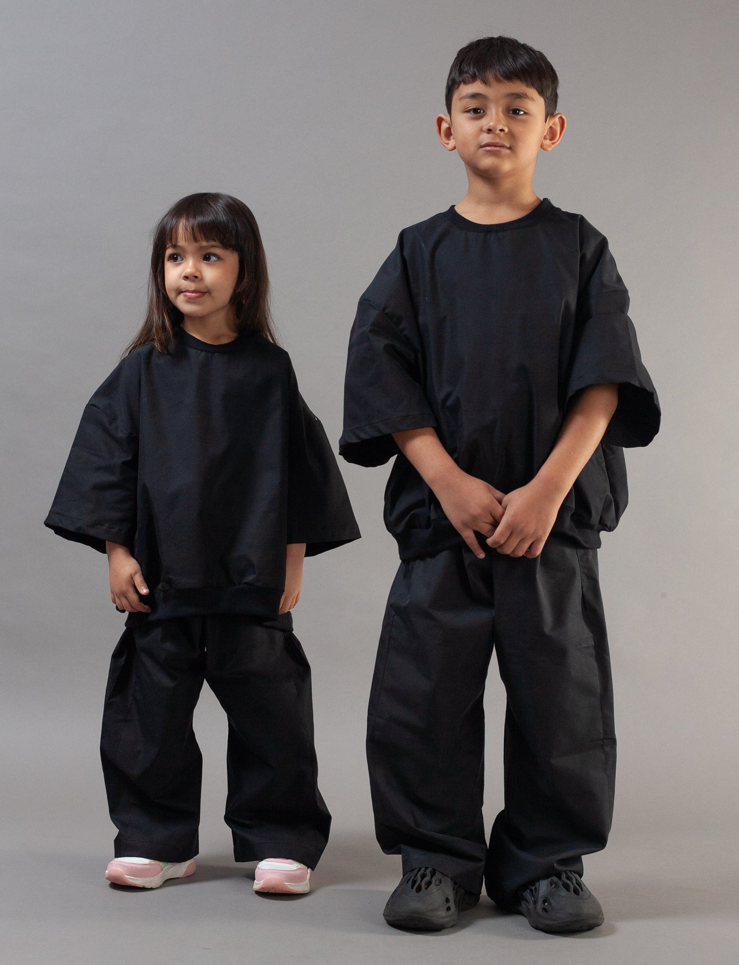 A young boy and a young girl modeling matching black wide-sleeve poncho-style outfits against a solid gray studio background.