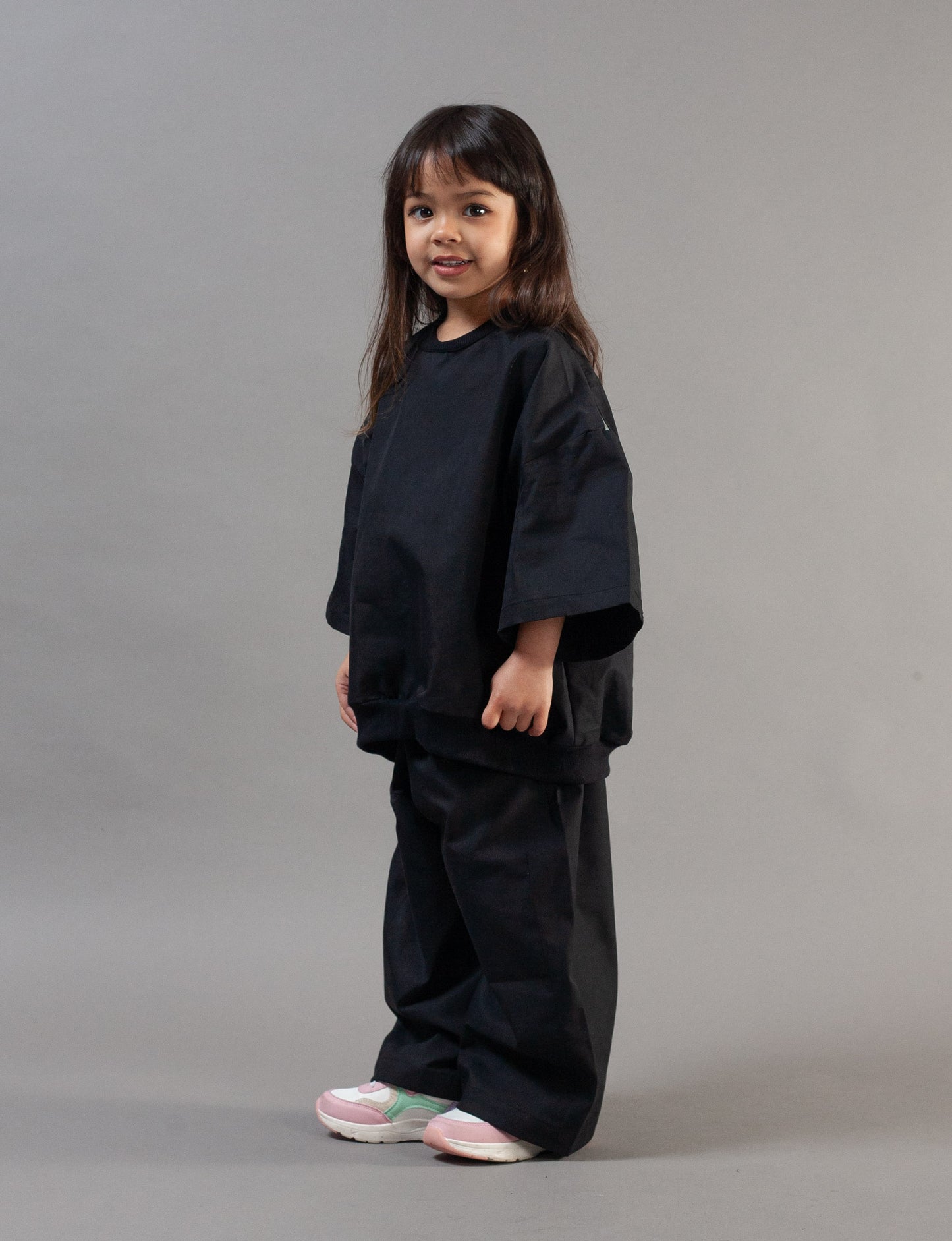 A young girl with long dark hair stands in profile wearing a black oversized short-sleeve top and matching black wide-leg pants, paired with light pink and white sneakers, against a solid gray studio background.