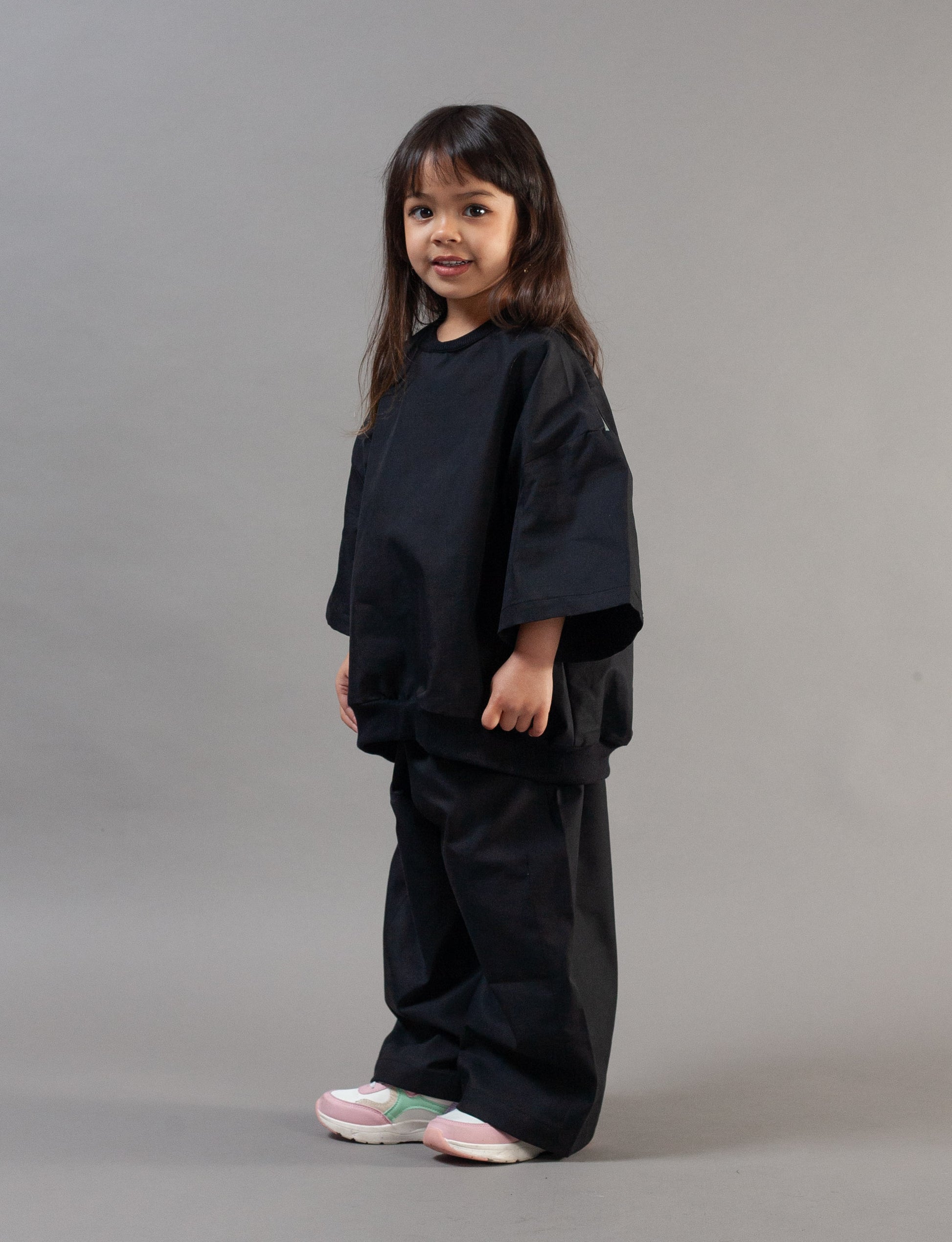 A young girl with long dark hair stands in profile wearing a black oversized short-sleeve top and matching black wide-leg pants, paired with light pink and white sneakers, against a solid gray studio background.