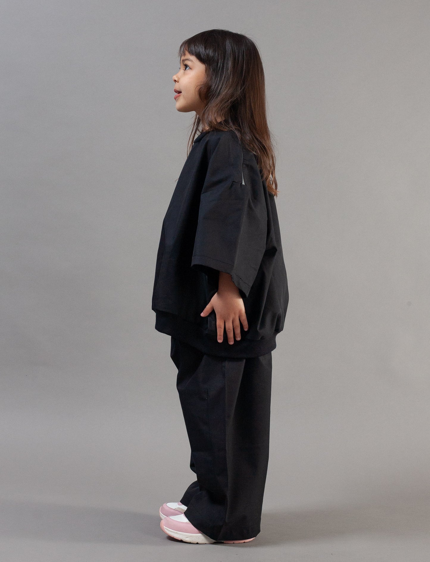 A young girl with long dark hair is shown in profile wearing a black wide-sleeved top and matching black pants, standing against a solid gray studio background.