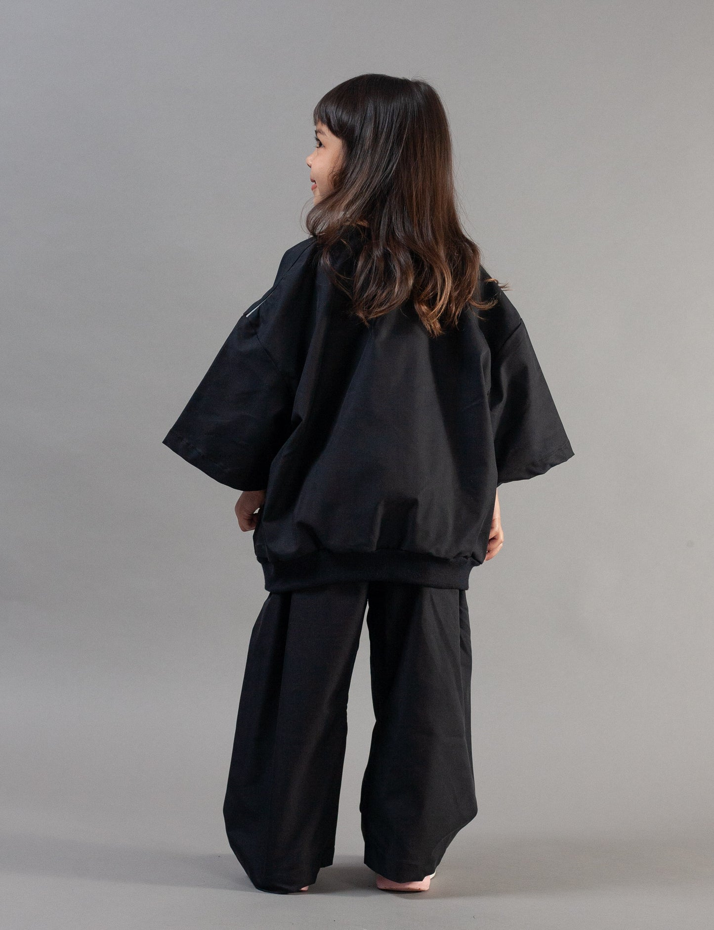 A child model shown from the back wearing a black wide-sleeved top and matching wide-leg pants against a plain gray studio background.