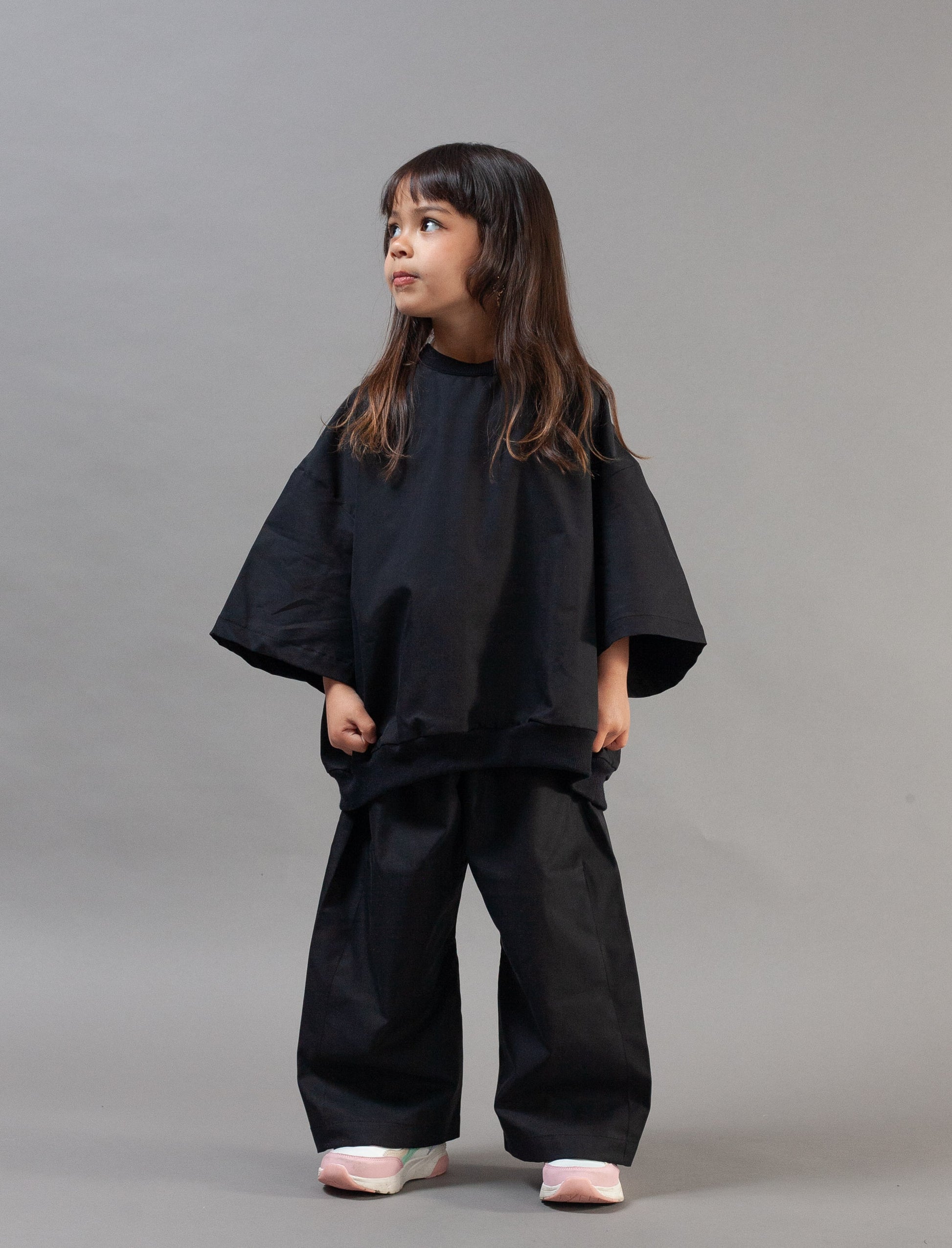A young girl with long dark hair stands against a solid gray studio background wearing a loose-fitting black two-piece outfit with wide sleeves and wide-leg pants, paired with light pink sneakers.