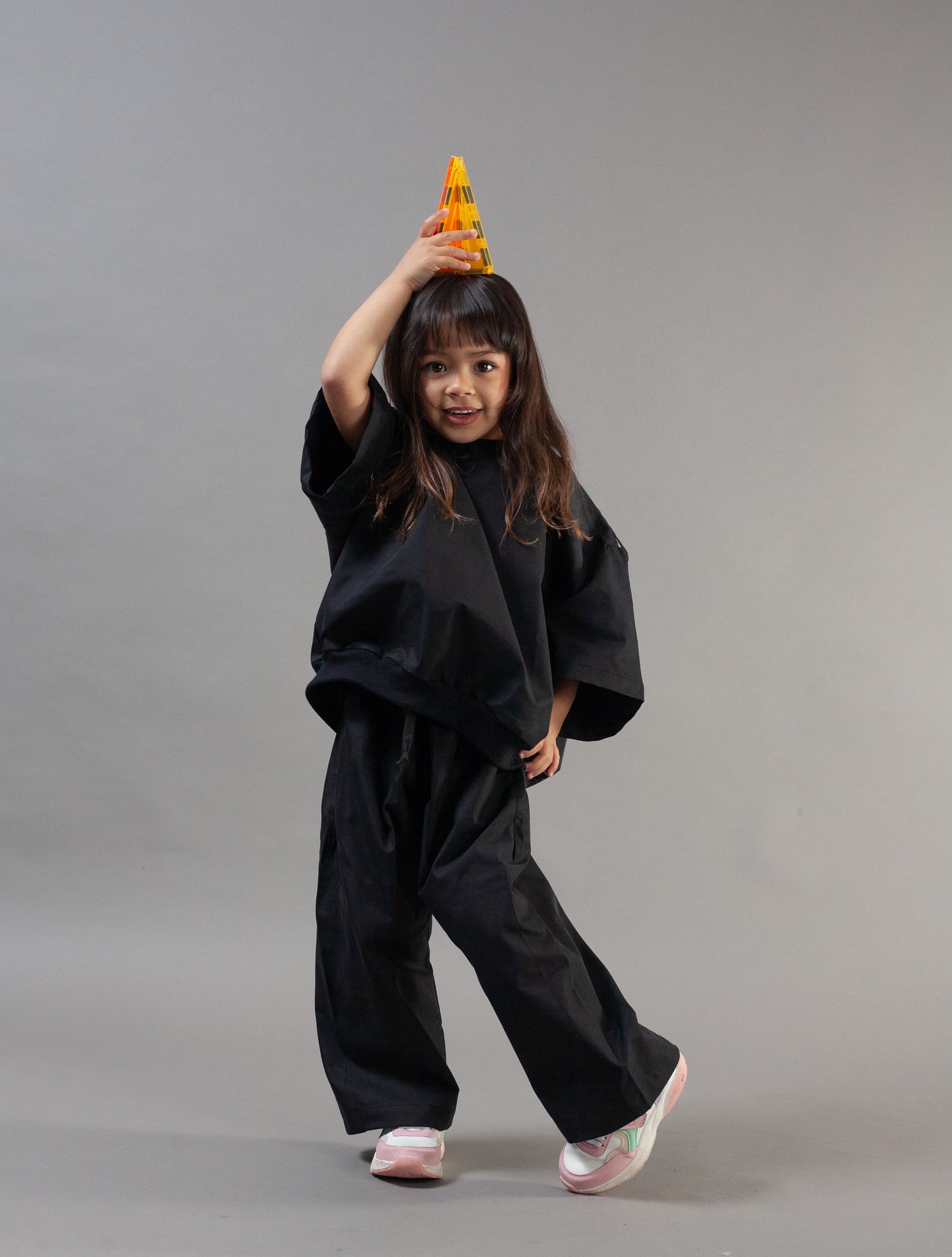 A young girl modeling a black wide-leg costume jumpsuit with a matching black headband, holding a small yellow party hat above her head, against a solid gray studio background.