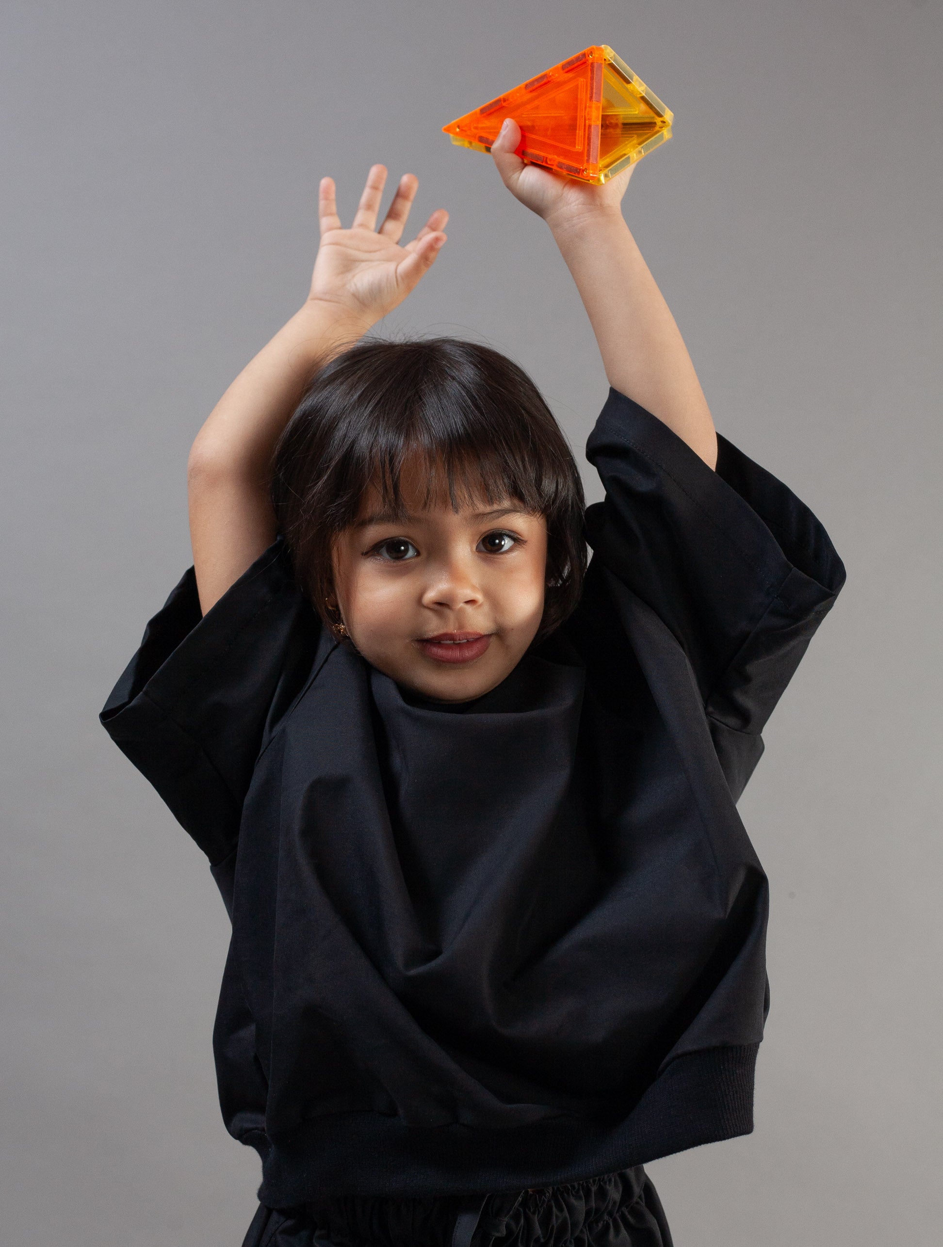 A young child wearing a black wide-sleeved top and face mask stands against a plain gray background, holding up a translucent orange and yellow geometric plastic toy with both arms raised.