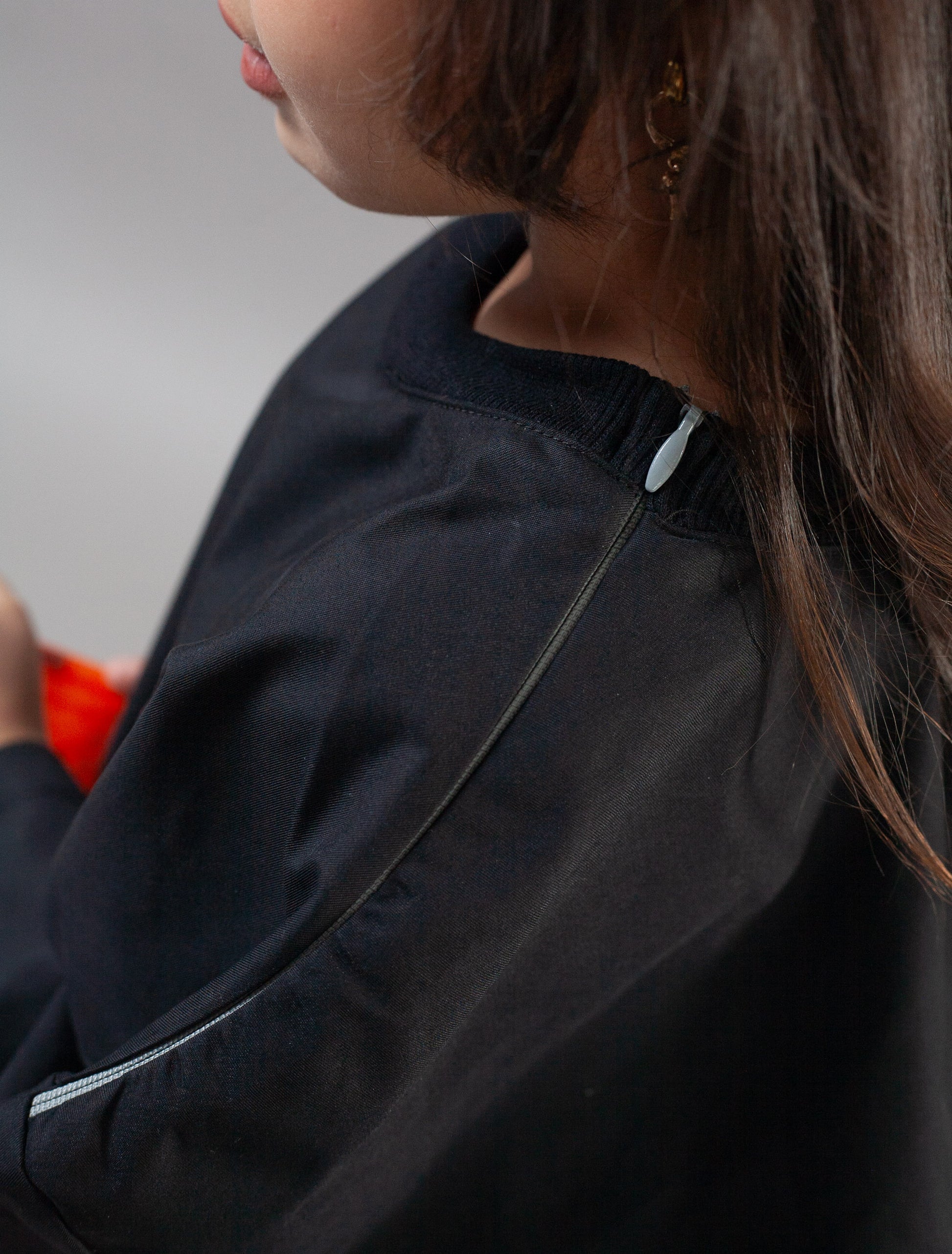 Close-up of a person wearing a dark navy or black zip-front jacket, showing part of the face, hair, shoulder, and sleeve against a plain light gray background.