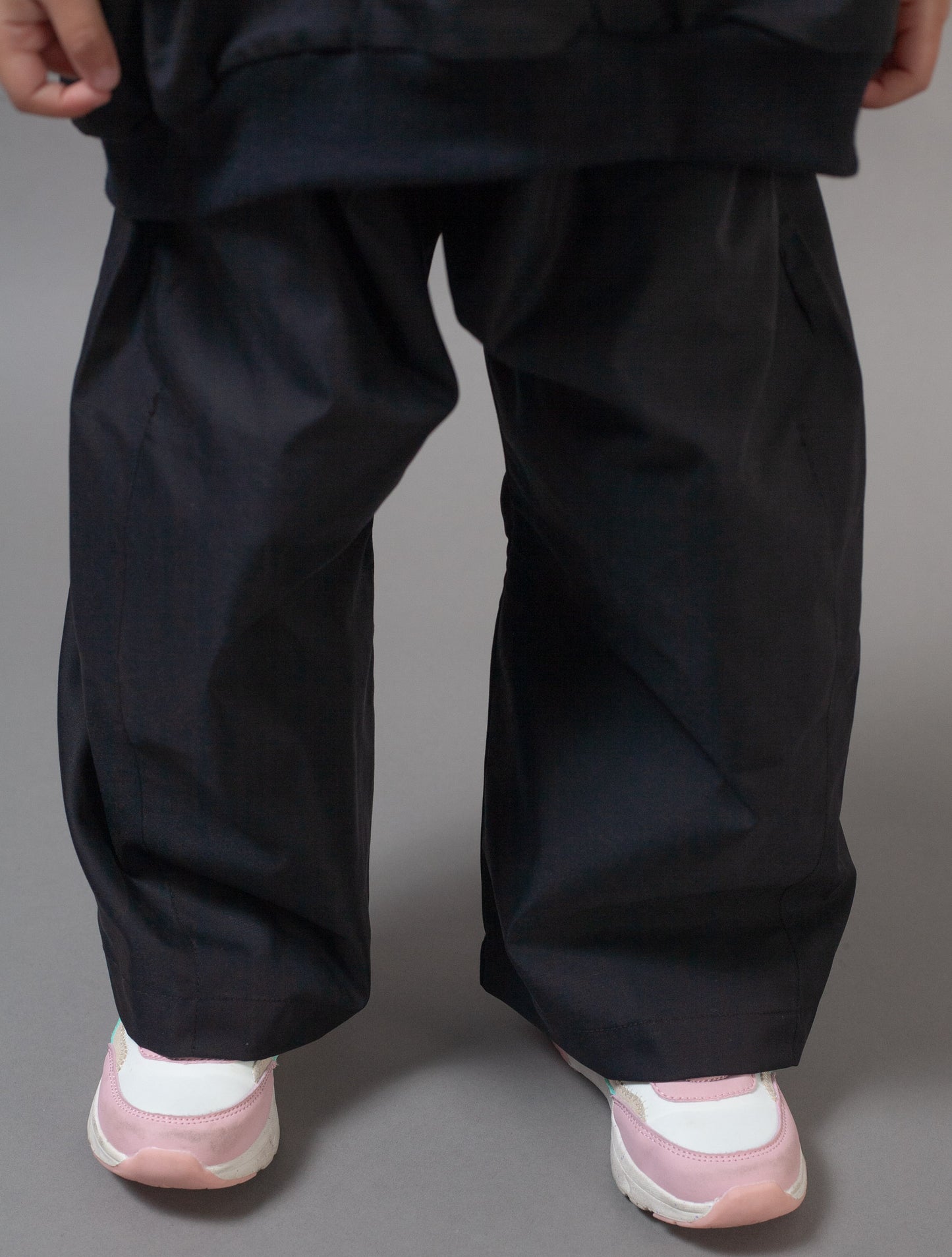 A close-up of a person's lower body wearing loose, oversized black pants and light pink and white sneakers against a solid gray studio background.