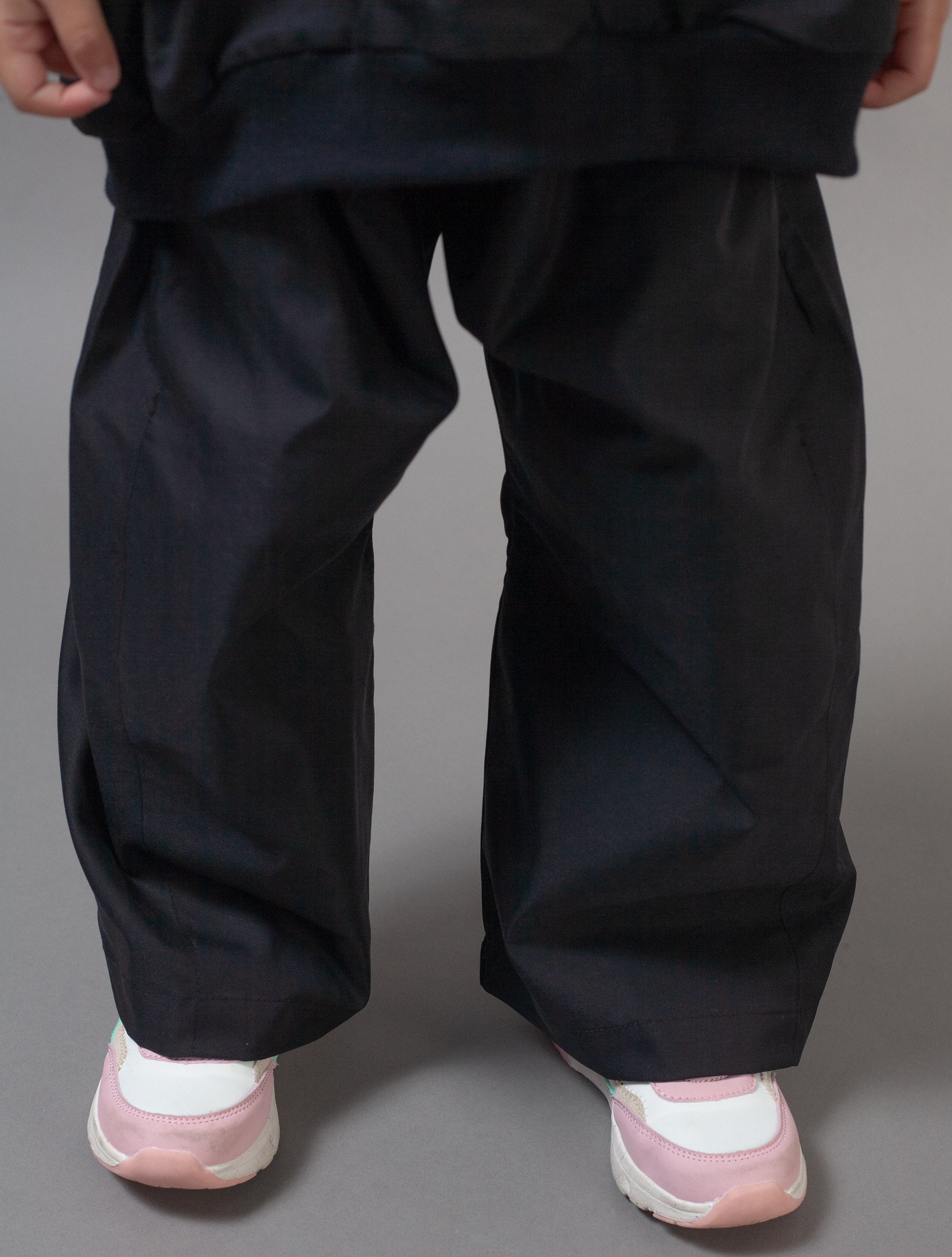 A close-up of a person's lower body wearing loose, oversized black pants and light pink and white sneakers against a solid gray studio background.