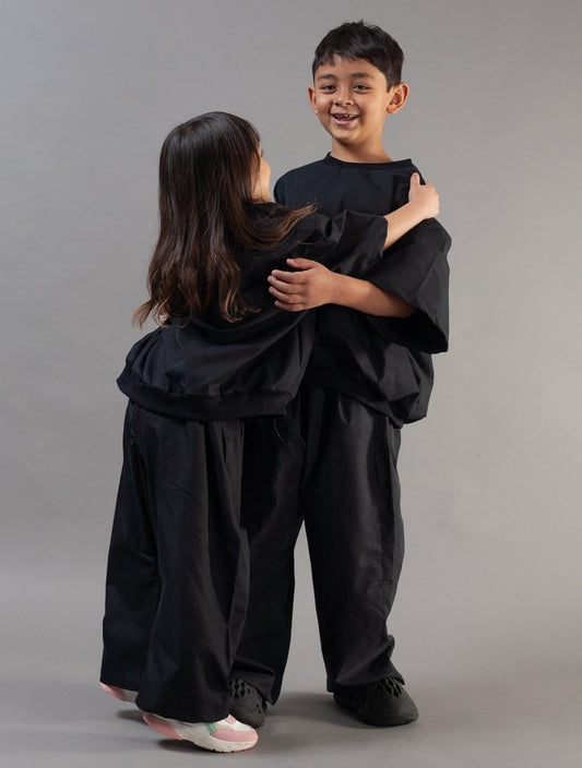 Two children wearing matching black martial arts uniforms embracing, photographed against a solid gray studio background.