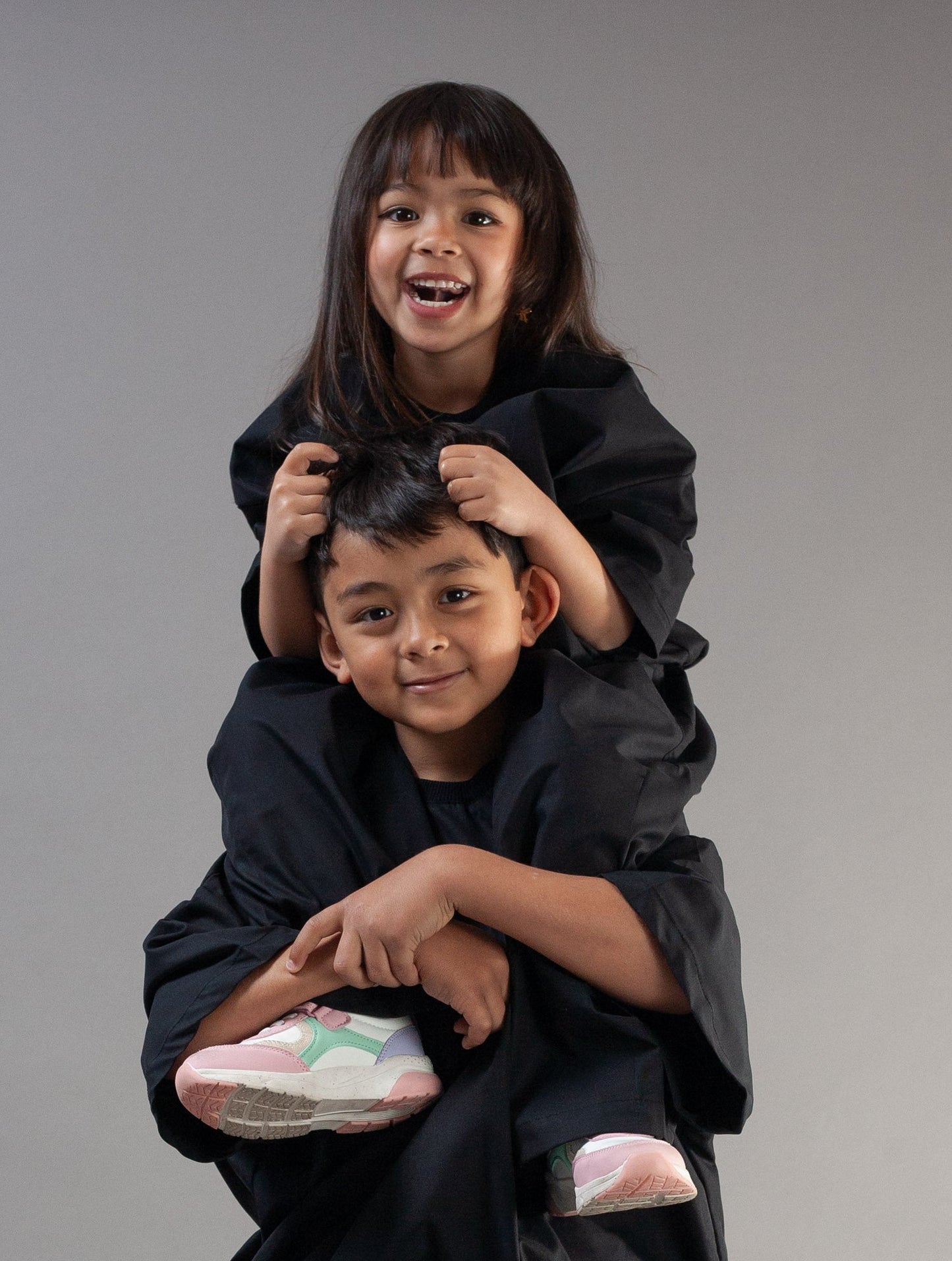A young boy wearing a black oversized jacket and matching pants carries a smiling girl on his shoulders, who is wearing a black top. They are posed against a solid gray studio background.
