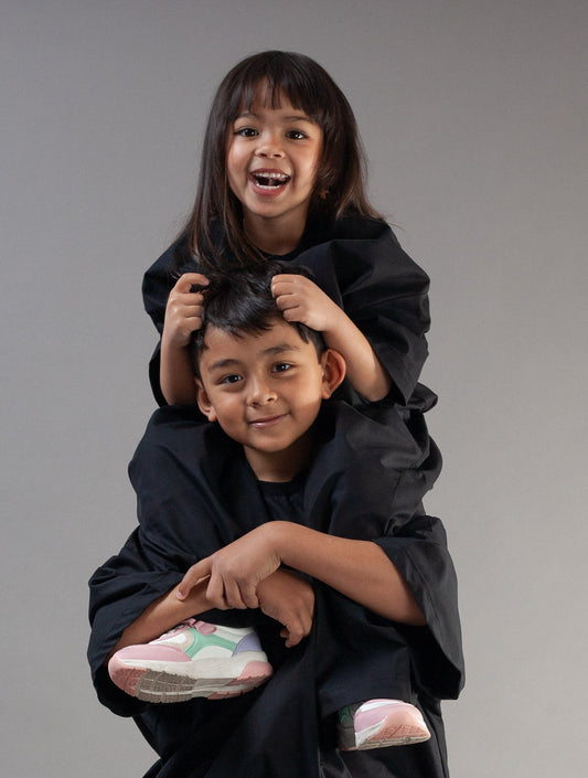 A young boy wearing a black oversized jacket and matching pants carries a smiling girl on his shoulders, who is wearing a black top. They are posed against a solid gray studio background.
