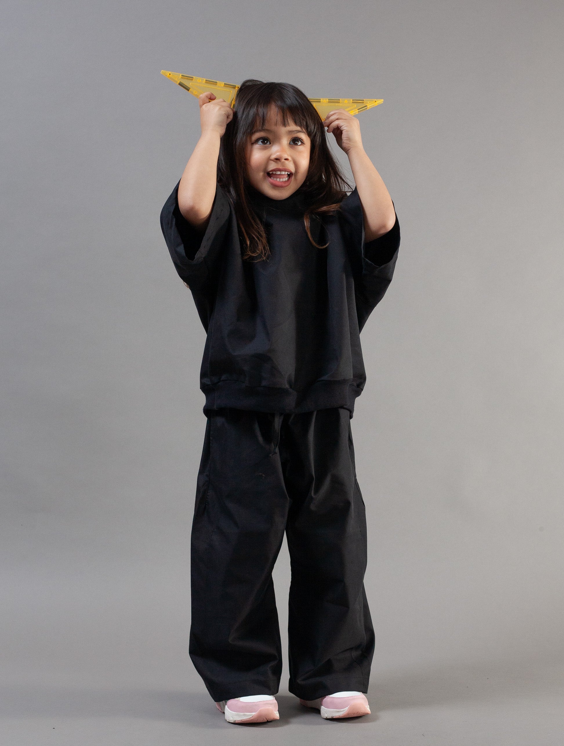 A young child wearing a black oversized sweatshirt and matching wide-leg pants, standing against a plain gray studio background and holding a yellow triangular object above their head.