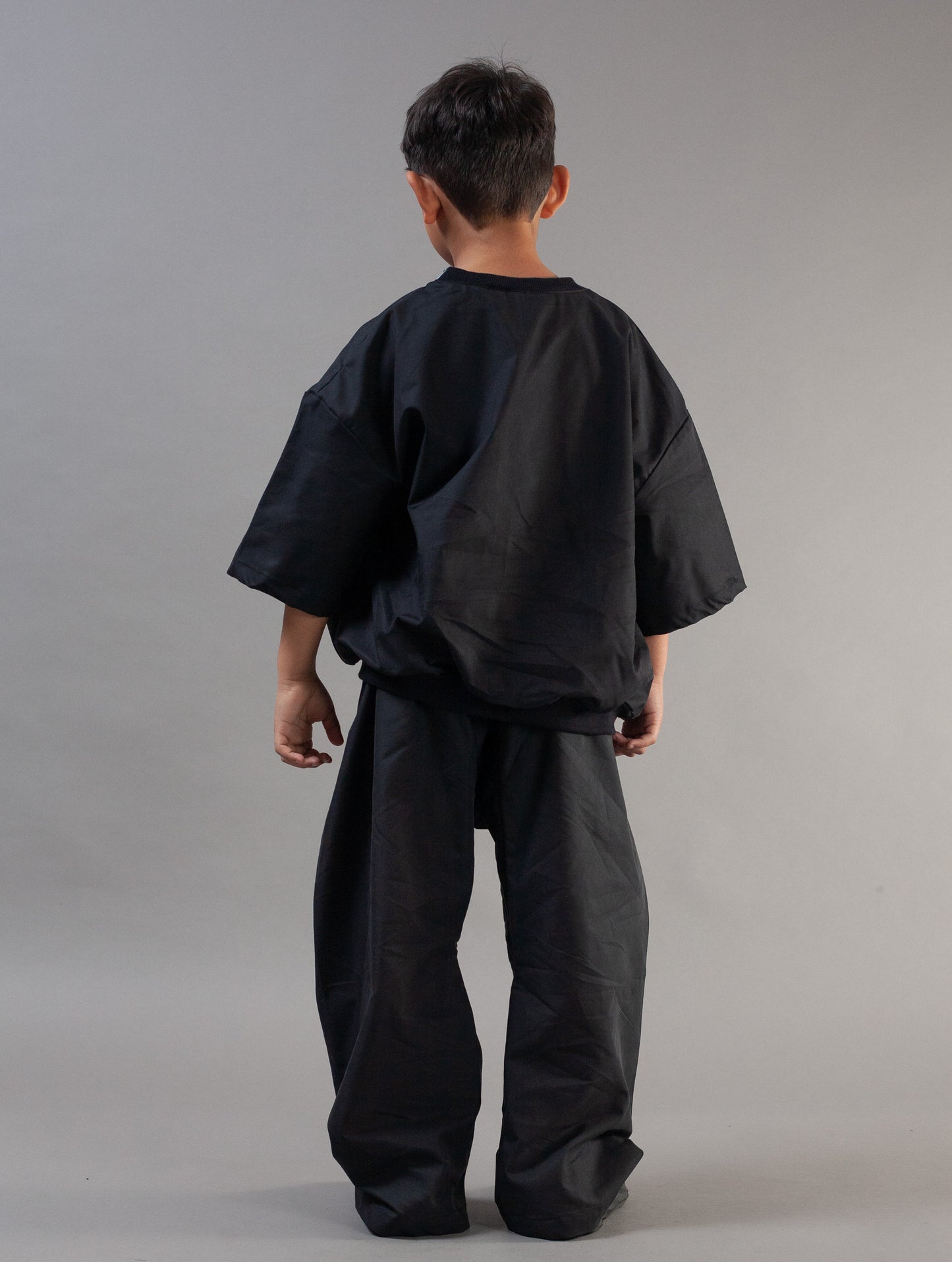 A child viewed from behind wearing an oversized black short-sleeve top and matching loose black pants against a plain light gray studio background.