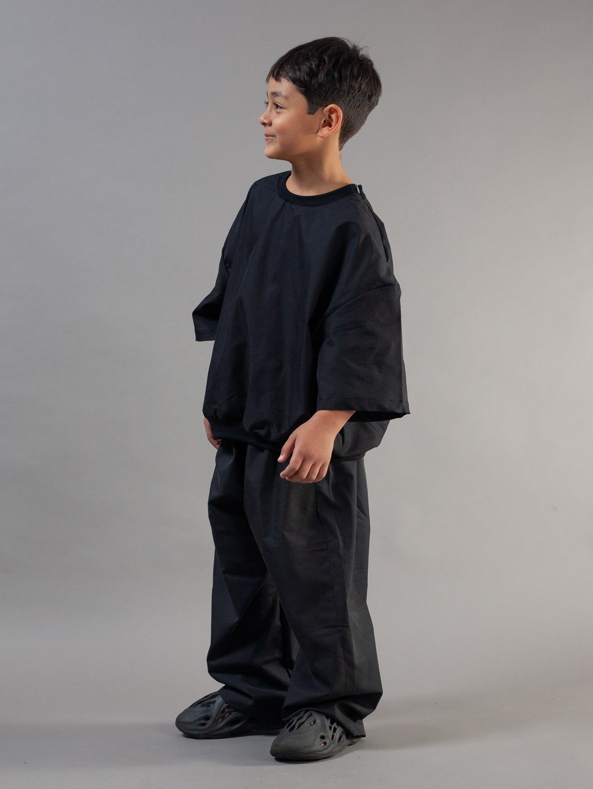 A young boy wearing an oversized black crewneck t-shirt and matching baggy black pants, standing in profile against a neutral gray studio background.